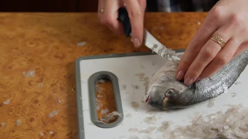 Hands scaling raw fish with knife on cutting board