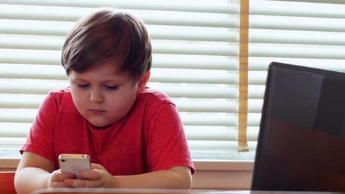Boy Using Phone at Table Beside Laptop