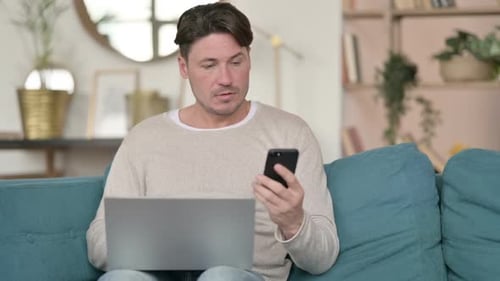 Mann mittleren Alters mit Smartphone arbeitet zu Hause am Laptop