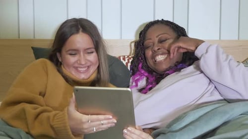 Close - Up Of Girlfriends Lying In Bed Using A Tablet Are At Home, Evening, Smiling, Cozy Apartment