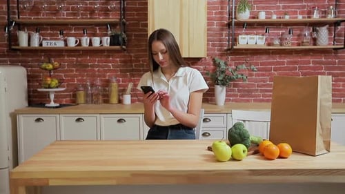 Woman Using Smartphone in Kitchen with Groceries