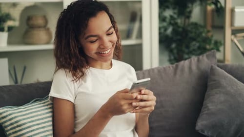 Joyful African American Woman Is Holding Smartphone, Touching Screen and Laughing Sitting on Couch