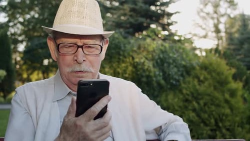 Portrait of Senior Man Holds Phone and Chats Thoughtfully on Web Camera in Park