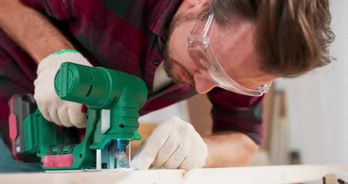 Der junge Zimmermann schneidet einen Holzbalken mit den Händen eines Puzzle-Handwerkers in Stoffschutzhandschuhen