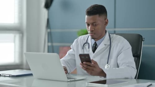 African Doctor using Smartphone while using Laptop in Office