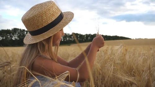 Happy Small Child in Straw Hat Holding Cereal Spikelet in Hands While Sitting in the Wheat Field