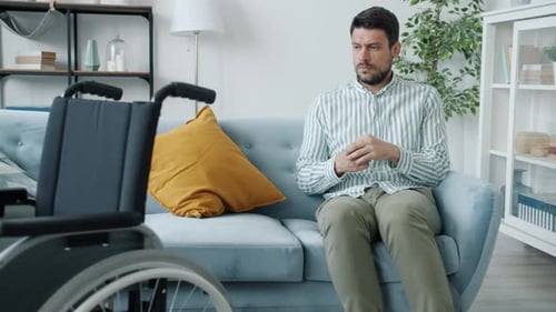 Man Sits Thoughtfully on Sofa Beside Wheelchair