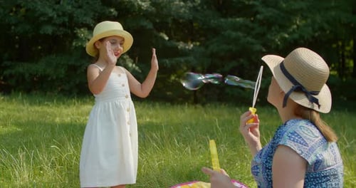 Funny Mom and Little Daughter Blow Soap Bubbles Sitting on the Lawn in the Park