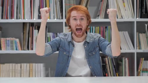 Man Cheering at Desk in Front of Bookshelf