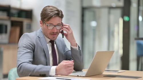 Businessman with Laptop Talking on Smartphone in Office