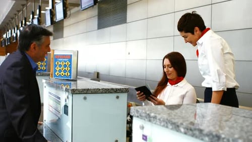 Two female airport staff checking passport and interacting with man at check-in desk