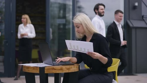Concentrated Female Analyst with Paperwork Analyzing Emarket on Laptop As Colleagues Passing at