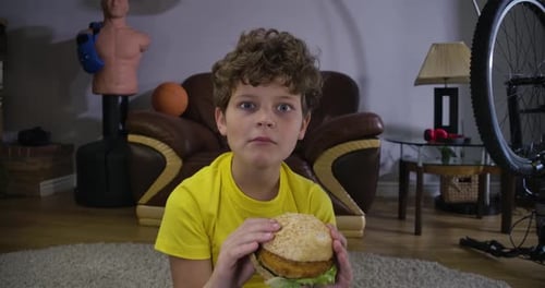 Visage d'un garçon caucasien aux cheveux bouclés mâchant un hamburger et regardant la télévision. Adolescent mignon avec restauration rapide
