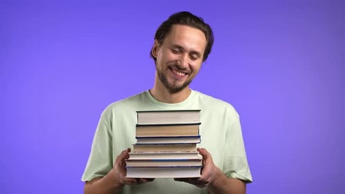 Male Student Holds Stack of University Books From Library on Violet Background in Studio