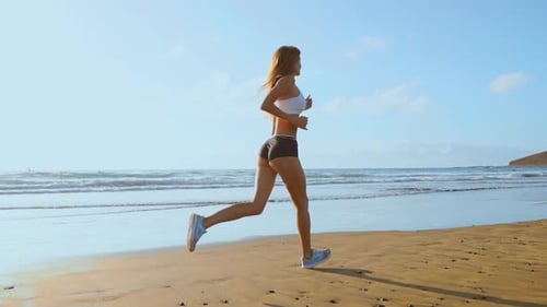 Beautiful Woman in Sports Shorts and T-shirt Running on the Beach with White Sand and Blue Ocean