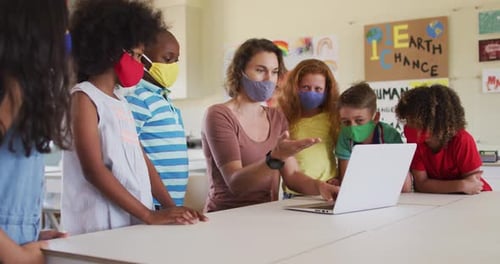 Female teacher wearing face mask using computer to teach kids in the class at school