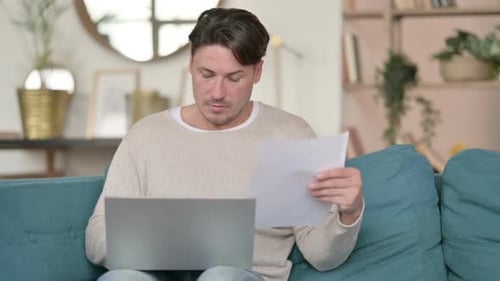 Man Works on Laptop With Papers at Home