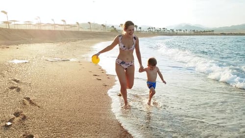 Footage of Happy Cheerful Mother with Toddler Son Running on the Sandy Sea Beach at Sunset