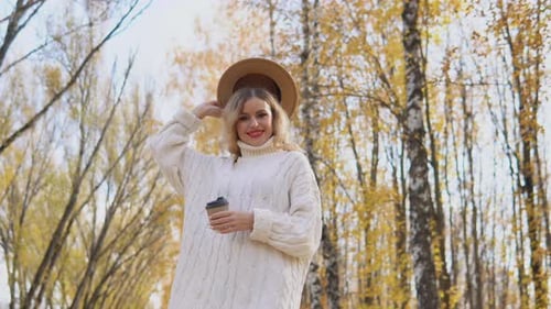 Beautiful Blonde Woman in a White Sweater Puts on a Brown Hat in the Autumn Park