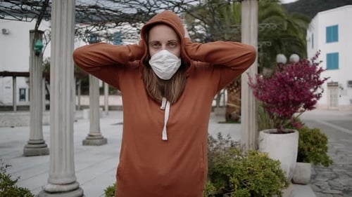Woman in orange sweater and medical mask stay and covers ears with hands on empty street