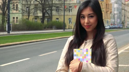 A Young Asian Woman Holds a Book Against Her Chest and Smiles at the Camera in a Street
