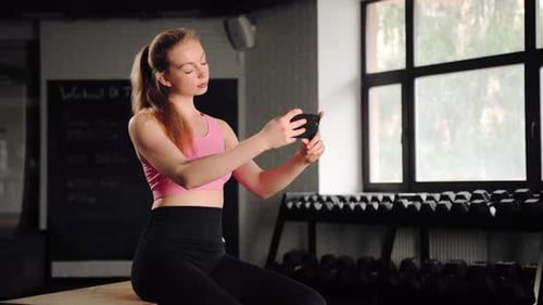 Athlete girl resting after exercise with the phone while sitting in gym.