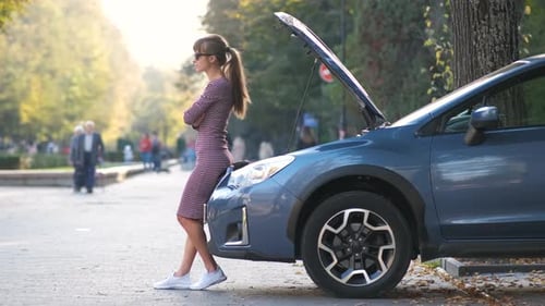Puzzled female driver standing on a city street near her car with popped up hood looking