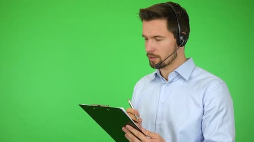 A Young Call Center Agent Talks To a Caller and Holds Notepad and a Pen, Then Smiles - Green Screen