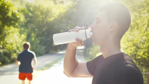 Side View Portrait of Young Man Jogger Drinking Water Resting During Run Training in Park in Morning