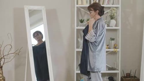 Vista lateral de uma elegante mulher sênior olhando no espelho experimentando um vestido azul pela manhã