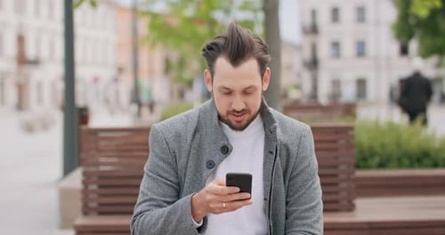 Young Man with Mustaches and a Beard Sitting on a Bench in the Square Scrolling the Smartphone