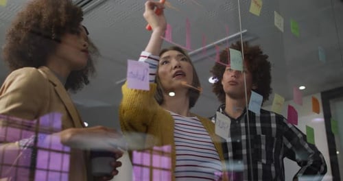 Des collègues de travail diversifiés utilisant des notes de service et écrivant sur un mur de verre