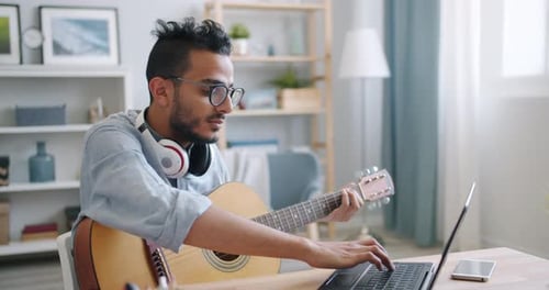Young Adult Plays Guitar and Types on Laptop
