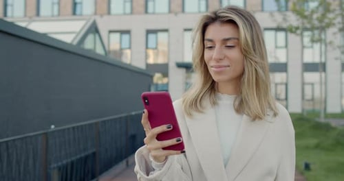 Attractive Millennial Woman with Blond Hair Using Smartphone While Walking at City Street. Crop View