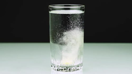 Eine sprudelnde gelbe Tablette gegen Schmerzen fällt in ein Glas und löst Zeitlupenaufnahme von