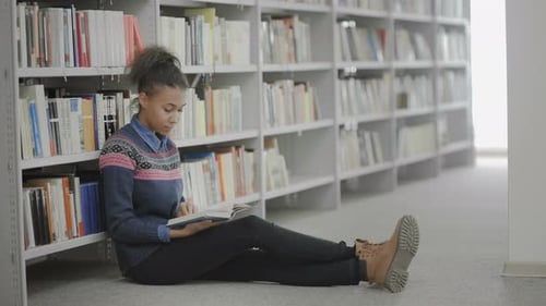 Young African American Woman Is Reading Book Sitting on Floor in School Library