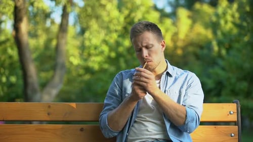 Jeune homme pensif fumant la cigarette, se relaxant sur le banc au parc, habitude nuisible