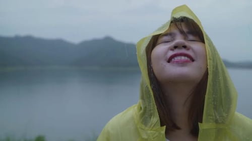 Young Asian woman feeling happy playing rain while wearing raincoat standing near lake.