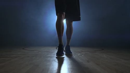 Männlicher Basketballspieler bewegt sich im Rauch vor der Kamera und klopft den Ball über den Parkettboden