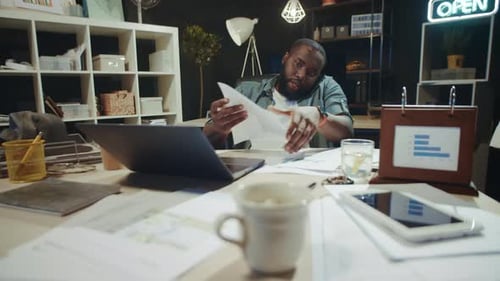 Portrait of African American Guy Working with Papers Near Laptop in Dark Office.