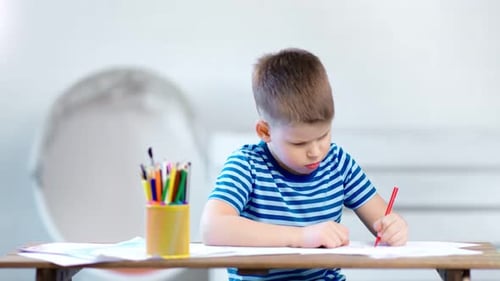Mittelgroße Aufnahme, selbstbewusster kleiner Junge, der mit Buntstiften ein Bild auf Papier zeichnet und auf dem Tisch sitzt