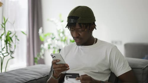 Young Black Man Holds Plastic Bank Card Sitting on a Comfortable Sofa Shopping Using Smartphone