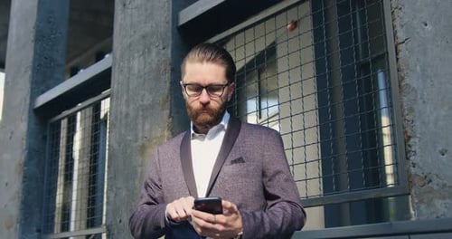 Hombre con gafas que espera a alguien mientras usa su teléfono cerca de la pared de un edificio moderno