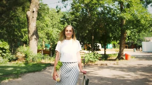Mujer joven en traje casual con comida en bolsas de compras orgánicas de malla está caminando en el parque de la ciudad