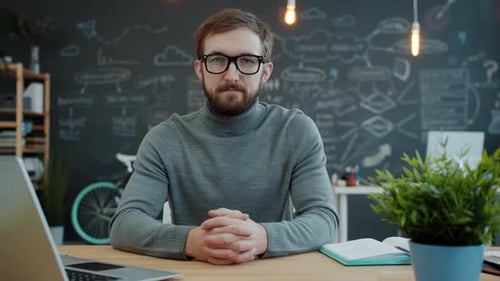 Portrait of Young Man in Glasses Looking at Camera Sitting at Desk, Laptop Computer Is Visible