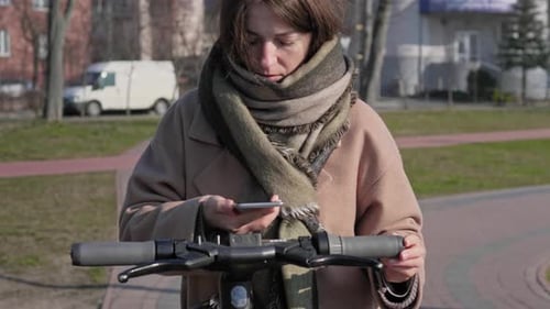 Woman Uses Phone with Electric Scooter in City Park