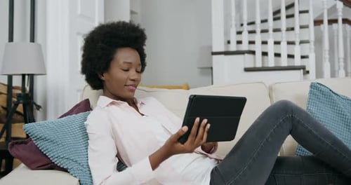 Mujer con peinado africano acostada en el sofá y disfrutando de la revisión de aplicaciones en una tableta