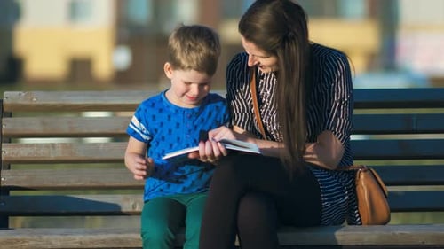 Young Mother and Son Sitting on a Bench in a Park and Reading a Book