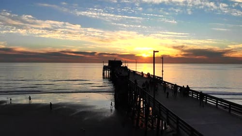 Drone shot rising over Newport Beach pier with many people walking in silhouette at sunset over the