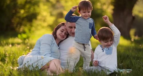 Dad Mom and Two Sons in the Summer at Sunset Sitting in a Meadow on the Grass Laughing and Hugging
